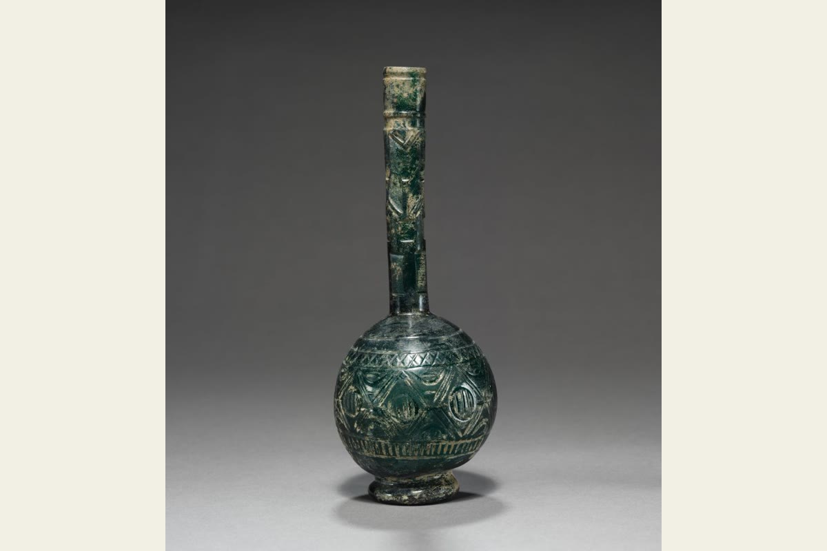 A dark blue-green glass bottle with a rounded body, long neck and carved geometric decoration.